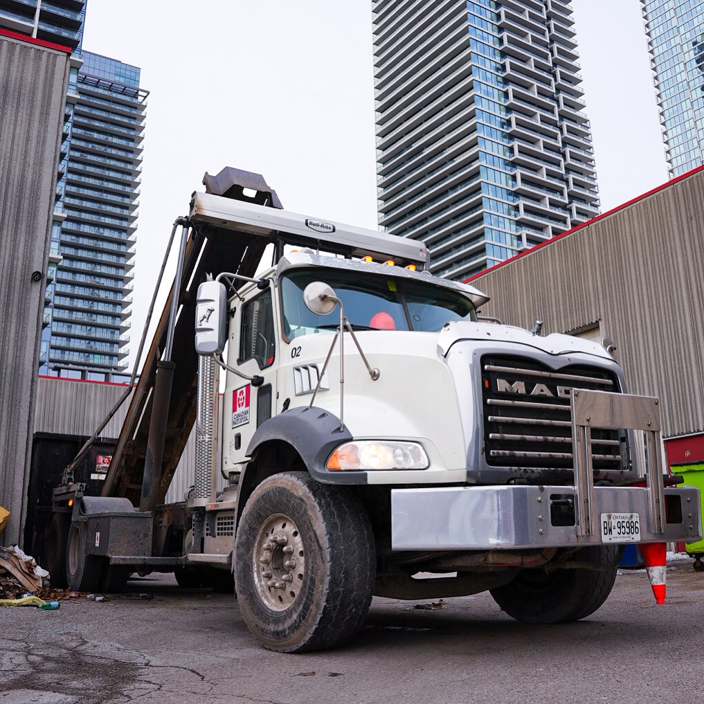All Canadian Waste Disposal A white Mack truck from All Canadian Waste Disposal is parked in an urban area near tall buildings and a construction cone, with waste bins and debris visible on the ground, highlighting local waste management efforts. residential and commercial waste management