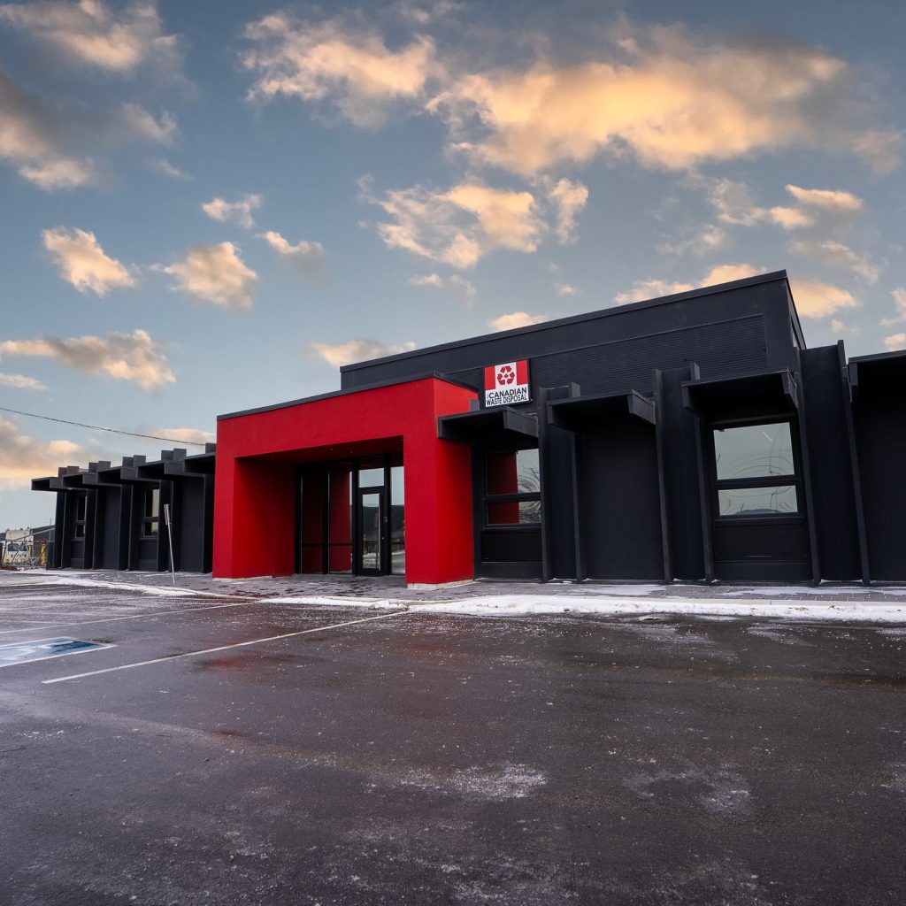 All Canadian Waste Disposal A modern commercial building with a red entrance and dark gray exterior sits under a cloudy sky, with an empty parking lot in front—ideal for businesses seeking convenient waste bins and reliable all Canadian waste disposal services for efficient waste management. residential and commercial waste management