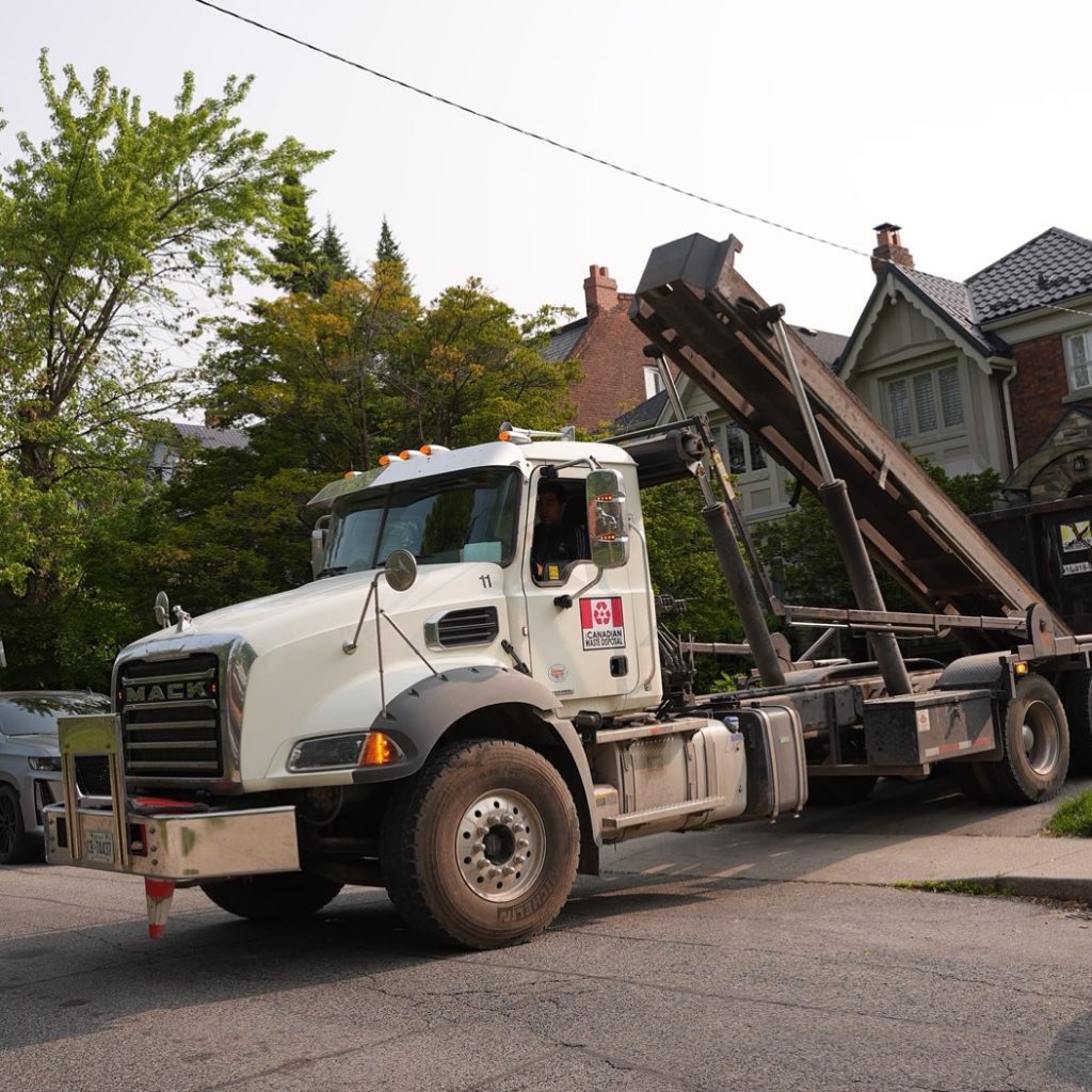 All Canadian Waste Disposal A white Mack truck with a tilt-frame bed, used for waste bins and waste management by All Canadian Waste Disposal, is parked on a residential street near houses and trees during the day. residential and commercial waste management